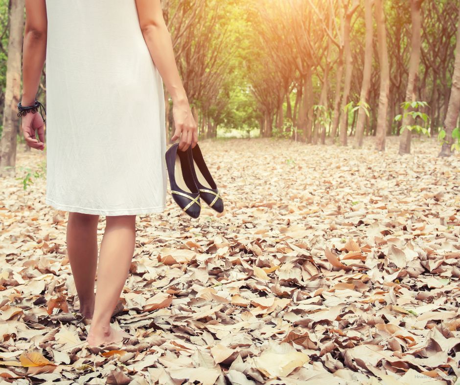 Woman walking barefoot on fallen leaves through a peaceful forest path, holding flats—capturing a grounding fall morning moment.