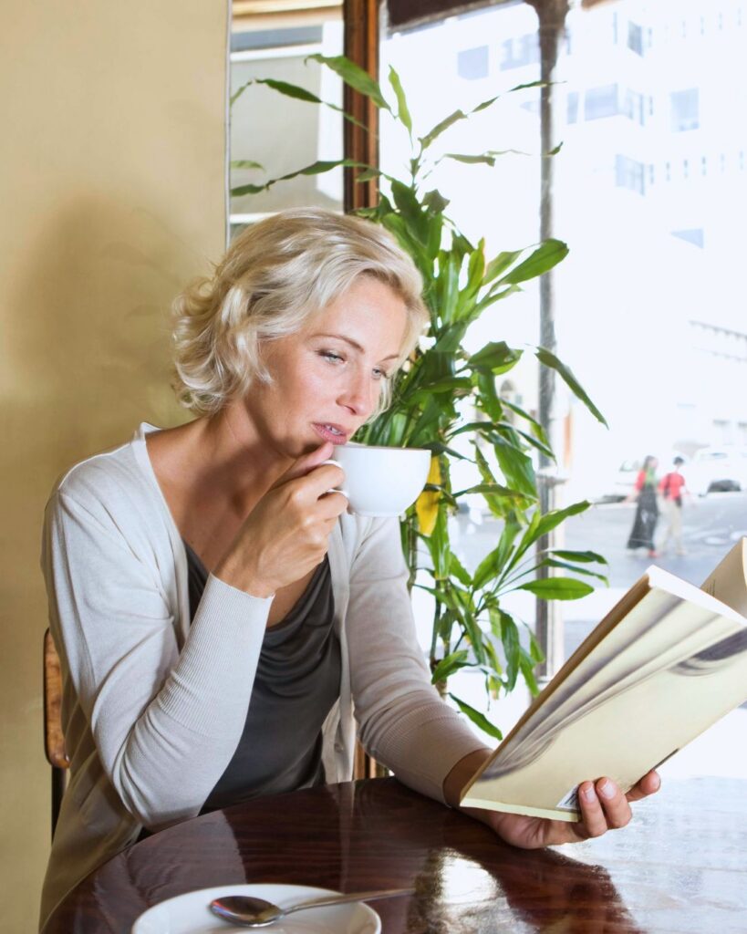 Woman reading a book and enjoying a cup of coffee by a sunny window, representing protected quiet time and making room for yourself in midlife.